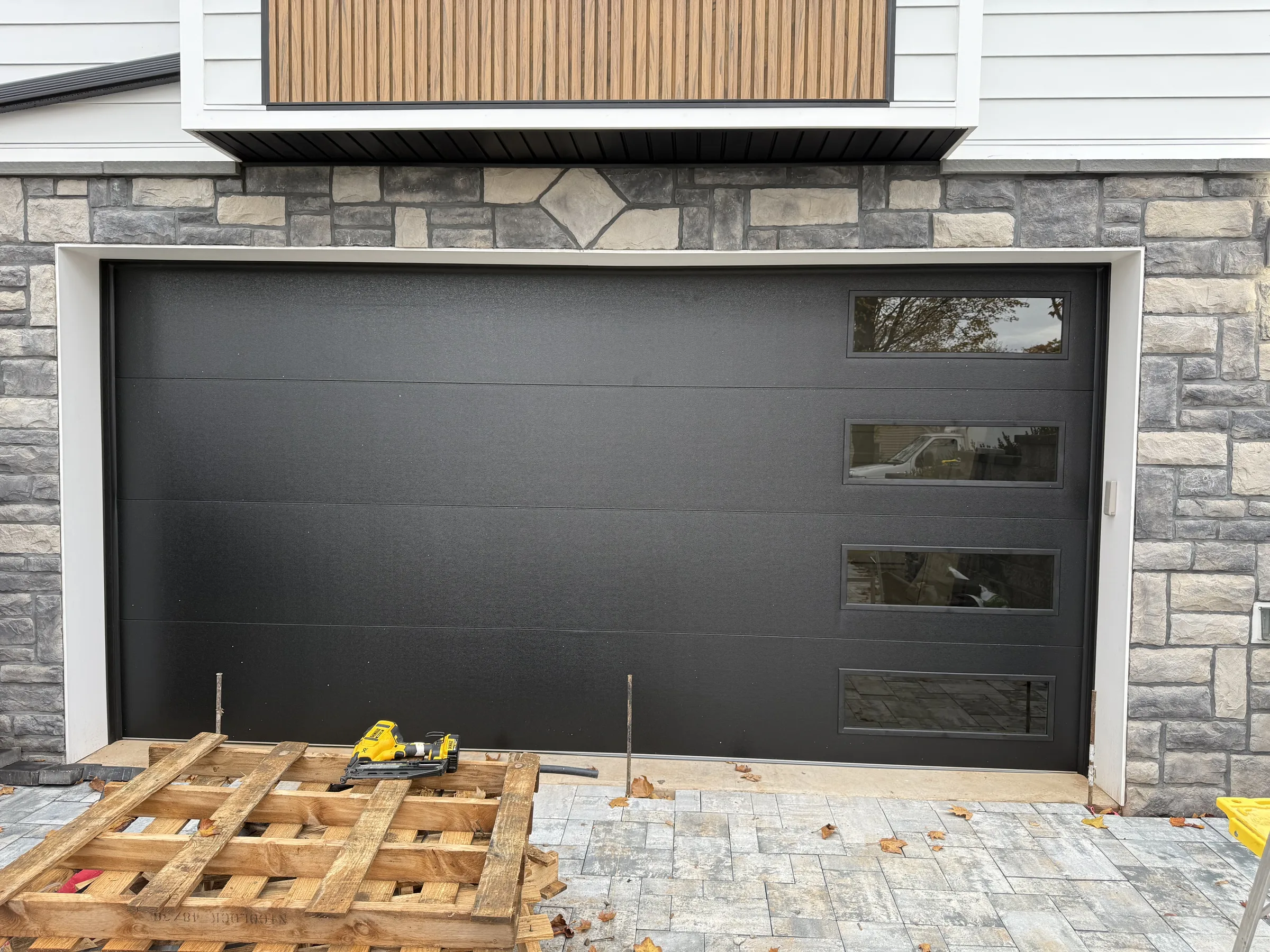 Black modern garage door with a horizontal side-window band installed on a stone-accented residential home.